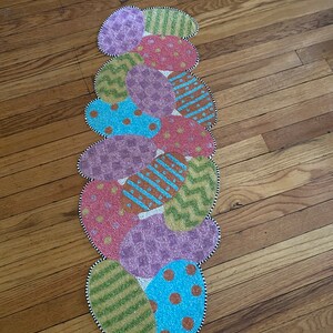 May include: A beaded table runner with a design of colorful Easter eggs. The runner is made of beads in shades of pink, purple, blue, green, and brown. The eggs are arranged in a vertical line, with the largest egg at the top and the smallest egg at the bottom.