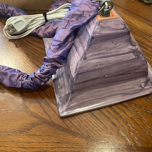 May include: A purple ceramic lamp base with a white cord and a purple fabric cord cover. The lamp base has a square shape with rounded edges and a textured surface.