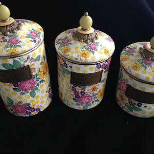 May include: Set of three decorative canisters with lids. The canisters feature a floral pattern with yellow polka dots, pink roses, yellow sunflowers, and purple flowers. Each canister has a brass-colored label and a yellow knob on top.