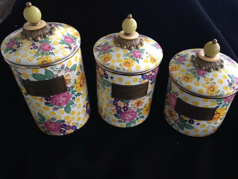 May include: Set of three decorative canisters with lids. The canisters feature a floral pattern with yellow polka dots, pink roses, yellow sunflowers, and purple flowers. Each canister has a brass-colored label and a yellow knob on top.