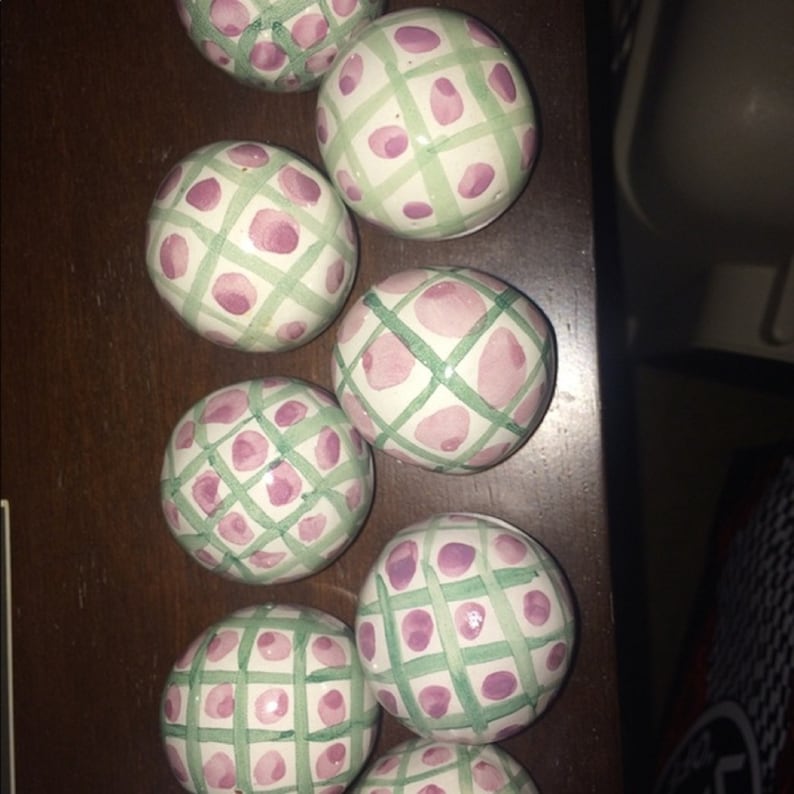 May include: Several white ceramic spheres with a hand-painted design. Each sphere has a light green grid pattern, with pink floral shapes at the intersections. The spheres are arranged on a dark brown surface.