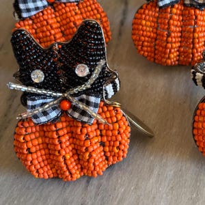 May include: A beaded cat-shaped ornament with a pumpkin-shaped body. The cat's head is black with rhinestone eyes and a black and white checkered bow tie. The pumpkin body is orange, and the ornament has a gold-colored metal tag.