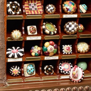 May include: A selection of decorative cabinet knobs in a wooden display case. The knobs feature diverse designs, including checkered, floral, and geometric patterns. The display case has brass nameplates. The knobs are in various colours.