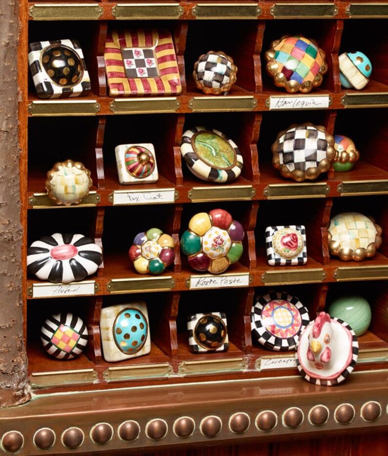 May include: A selection of decorative cabinet knobs in a wooden display case. The knobs feature diverse designs, including checkered, floral, and geometric patterns. The display case has brass nameplates. The knobs are in various colours.