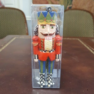 May include: A colorful nutcracker ornament in a clear plastic box. The nutcracker is wearing a red and white checkered jacket, blue and green pants, and a blue and green crown. The nutcracker has a white face with a black mustache and a black beard.