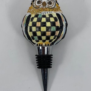 May include: A decorative bottle stopper featuring an owl design. The owl has a black and white head, gold glitter accents, and a checkered body in black, yellow, and green. The stopper has a silver metal base and a black rubber seal.