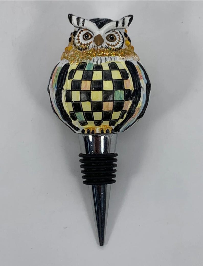 May include: A decorative bottle stopper featuring an owl design. The owl has a black and white head, gold glitter accents, and a checkered body in black, yellow, and green. The stopper has a silver metal base and a black rubber seal.