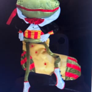 May include: A green frog wearing a tuxedo and a yellow stocking with red and green stripes. The frog is holding a red ribbon in its hand.