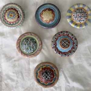 May include: Six decorative ceramic knobs with detailed patterns. The knobs display various designs, including floral and geometric patterns, in colours such as blue, yellow, red, and green. Each round knob has a unique, hand-painted appearance. Measurements are in centimetres.
