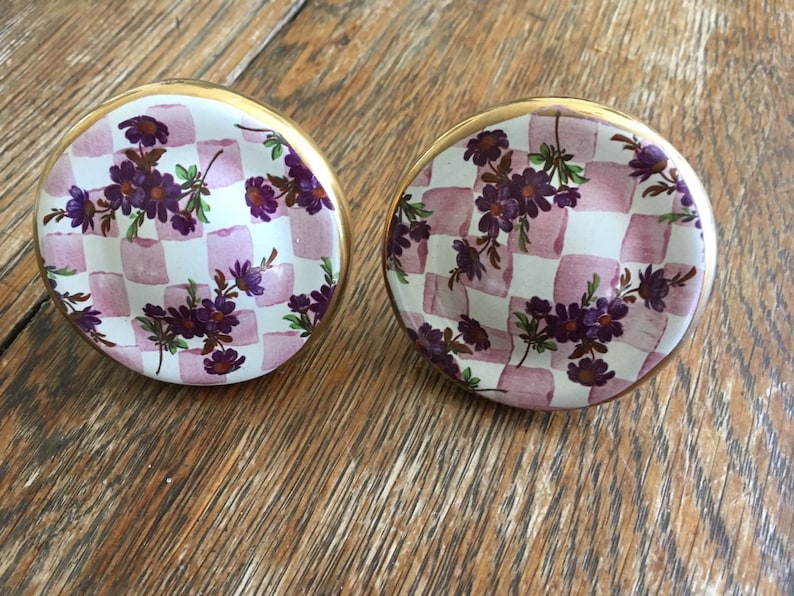 Puede incluir: Dos pomos de armario redondos de cer&aacute;mica con borde dorado. Los pomos presentan un patr&oacute;n de cuadros en blanco y rosa claro, adornados con dise&ntilde;os florales morados. Estos pomos decorativos son ideales para a&ntilde;adir un toque de elegancia a los muebles.