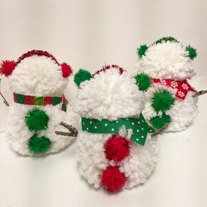 May include: Three white yarn pom-pom snowmen ornaments with red and green accents. The snowmen have red pom-poms for noses, green pom-pom hats, and green and red ribbon accents.