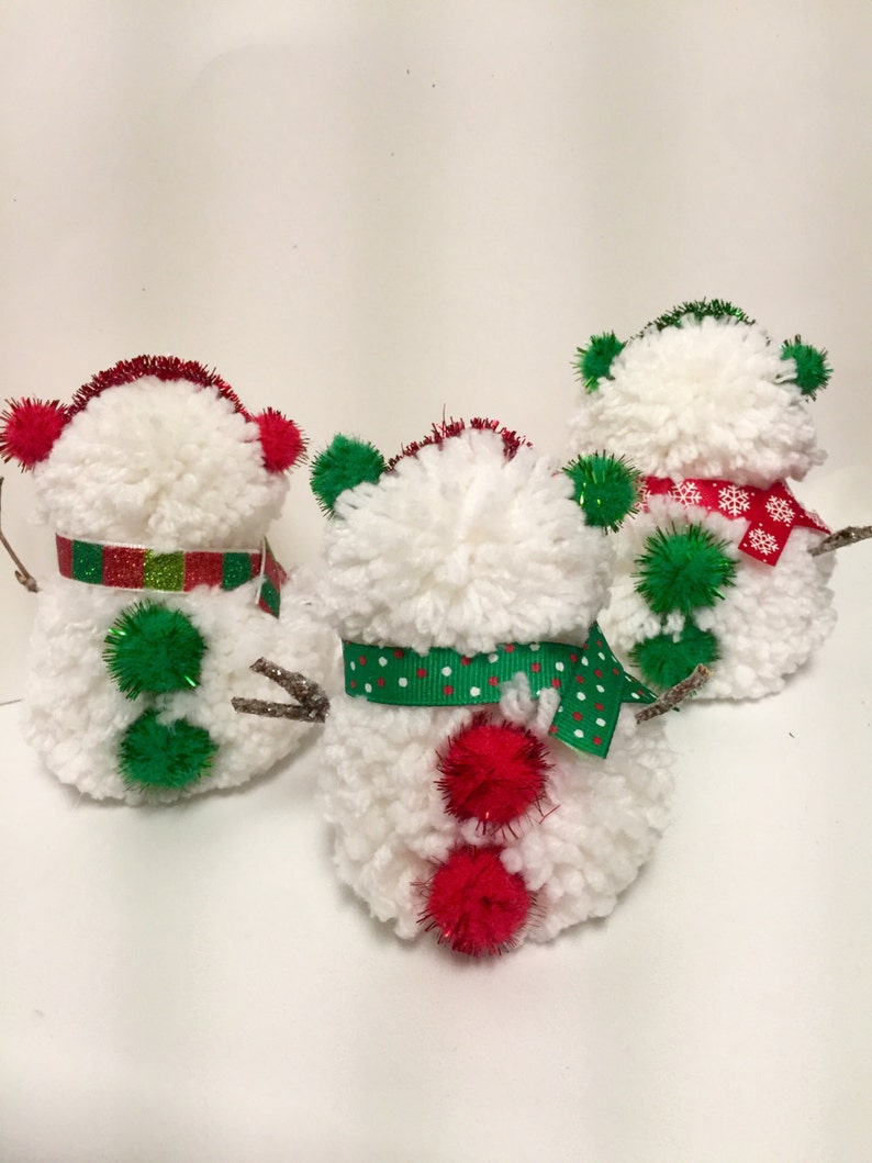 May include: Three white yarn pom-pom snowmen ornaments with red and green accents. The snowmen have red pom-poms for noses, green pom-pom hats, and green and red ribbon accents.