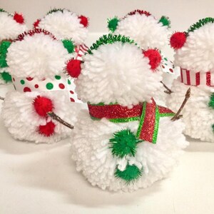 May include: A group of four white yarn pom-pom snowmen with red and green accents. Each snowman has a different design, including a red and white striped scarf, a red and green polka dot scarf, a red and green ribbon, and a green pom-pom hat.