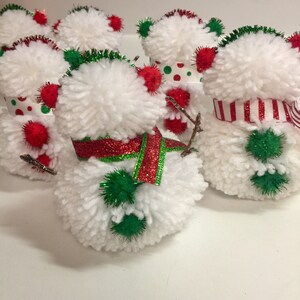 May include: Five white yarn pom-pom snowmen with red and green accents. The snowmen are decorated with red and white striped ribbon, red glitter ribbon, and green pom-poms.