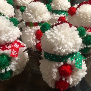 May include: A group of five white yarn pom-pom snowmen ornaments with red and green accents. Each snowman has a red pom-pom hat, a green pom-pom scarf, and a red pom-pom nose.