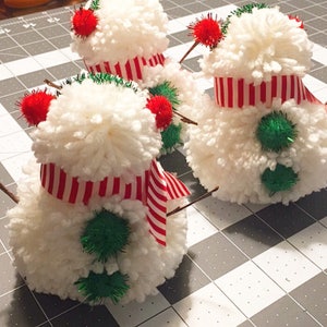 May include: Three white yarn pom-pom snowmen with red and white striped ribbon scarves and green pom-poms for buttons.