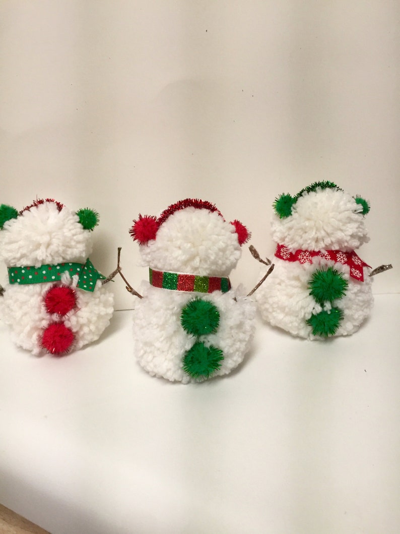 May include: Three white yarn pom-pom snowmen with green and red accents. The snowmen are decorated with green and red pom-poms, ribbon, and snowflake patterned fabric.