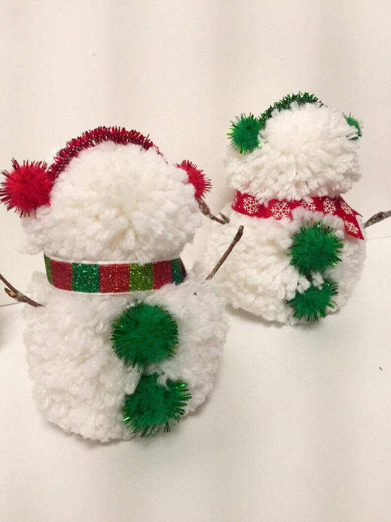 May include: Two white pom-pom snowmen with green and red accents. One snowman has a red and green striped scarf and green pom-poms for buttons. The other snowman has a red scarf with white snowflakes and a green pom-pom on the back.