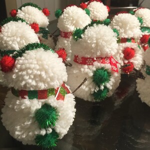 May include: A group of white pom-pom snowmen with red and green accents. The snowmen are decorated with red and white striped scarves, green pom-poms, and red and white snowflake patterns.