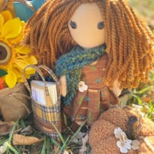 May include: A handmade fabric doll with brown yarn hair, wearing a brown and white dress with a green scarf, holding a small plaid bag. The doll is standing next to a small brown pumpkin with white flowers.