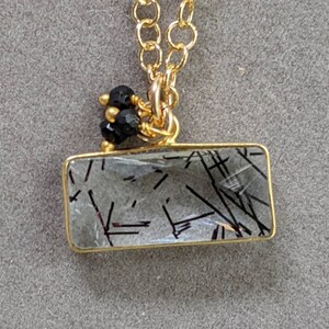 May include: A gold chain necklace with a clear quartz pendant featuring black inclusions. The pendant is rectangular and has a gold frame. The chain has a small cluster of black beads at the top.