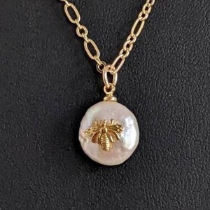 12mm coin pearl enhanced with a gold bee hangs on a sealed gold-plated brass Figaro chain, adjustable via a lobster clasp with seed pearls