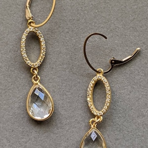 May include: Gold dangle earrings with clear teardrop-shaped crystals and oval-shaped frames with clear stones.