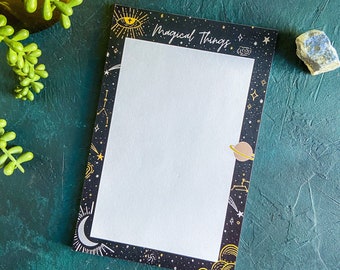 May include: A black notepad with a white border and gold accents. The border features a celestial design with stars, planets, and a crescent moon. The notepad has the text "Magical Things" written in gold lettering.