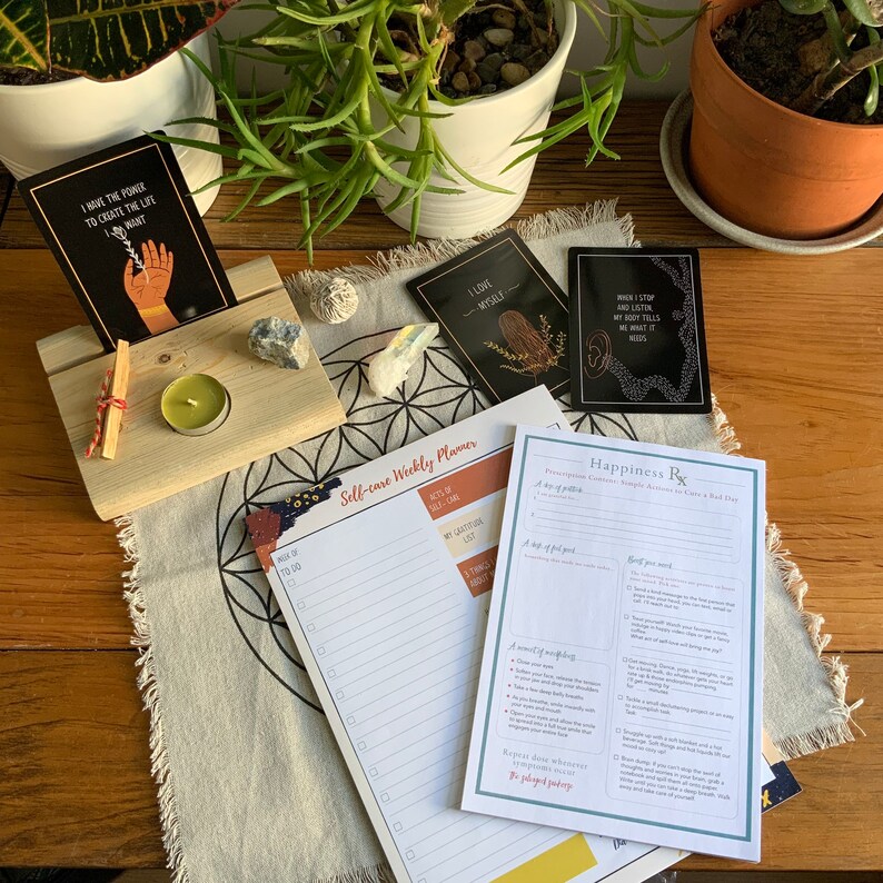 May include: A self-care weekly planner and a happiness prescription card with a list of suggestions for a bad day. The planner has a section for acts of self-care, a gratitude list, and a to-do list. The prescription card includes suggestions for a cup of tea, a nap, a good meal, and a moment of mindfulness. The cards are on a table with a wooden stand, a candle, and crystals.