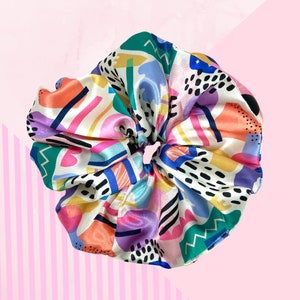 May include: A hair scrunchie featuring a smooth, satin-like fabric with a vibrant, multi-coloured abstract pattern. The design includes geometric shapes, zig-zag lines, and black dotted accents in shades of pink, blue, yellow, orange, and green.