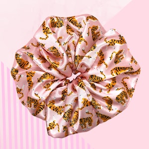 May include: A large, pink satin scrunchie featuring a repeating pattern of orange and black tigers. The scrunchie is gathered in the centre and has a shiny, smooth texture. The background is light pink with vertical stripes.