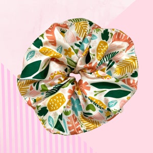May include: A large, white satin scrunchie with a vibrant botanical print. The design features green, yellow, and orange leaves, flowers, and other plant elements. The scrunchie is designed for hair and is set against a pink background.