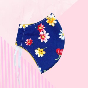 Puede incluir: Una mascarilla azul marino con un estampado floral de flores rojas, amarillas y blancas. La mascarilla tiene cintas elásticas blancas para las orejas y un forro amarillo. El fondo es un degradado de rayas rosas y blancas.