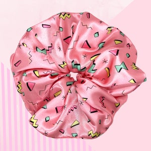 May include: A large, pink satin scrunchie with a retro geometric pattern. The design includes shapes like triangles, zigzags, and stars in shades of pink, yellow, green, and black. The scrunchie is a hair accessory.