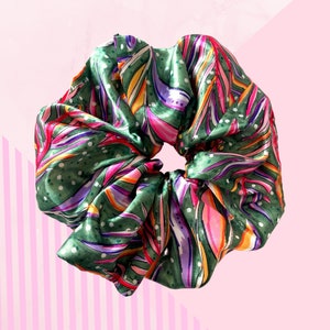 May include: A large, green satin scrunchie with a vibrant, abstract floral pattern in pink, purple, orange, and yellow. The scrunchie has small white polka dots and is set against a pink background.