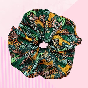 May include: A large, dark green scrunchie featuring a pattern of orange leopards, flowers, and foliage. The hair accessory is made of a soft, silky fabric. The scrunchie is designed to hold hair in a ponytail or bun.
