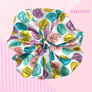 May include: A white satin scrunchie featuring a repeating pattern of colourful candy heart shapes. Each heart has a different phrase, such as "zero fucks given" and "you're fucking awesome". The word "EXPLICIT!" is in the upper right corner.