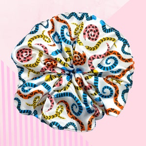 May include: A white satin hair bonnet featuring a vibrant snake print. The snakes are depicted in shades of blue, orange, yellow, and pink, set against a white backdrop. The bonnet is gathered at the top.