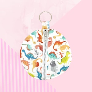Op de afbeelding: Een ronde, witte etui met ritssluiting en een kleurrijk dinosauruspatroon. De dinosaurussen zijn in tinten oranje, blauw, groen en geel. Een zilveren ritssluiting loopt in het midden en een zilveren sleutelhanger is aan de bovenkant bevestigd.