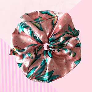 May include: A large, pink satin scrunchie with a floral design. It features teal and green leaves with pink accents and white polka dots. The background is a light pink with a striped pattern.