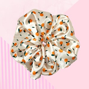 May include: Cream satin scrunchie featuring a pattern of orange fruit and green leaves. This oversized hair accessory is a stylish addition to any hairstyle. The scrunchie is a fun and fashionable hair accessory.