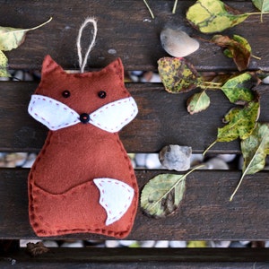 May include: A brown felt fox ornament with white felt accents for the ears, nose, and tail. The fox is hanging from a string and is on a wooden surface with autumn leaves.