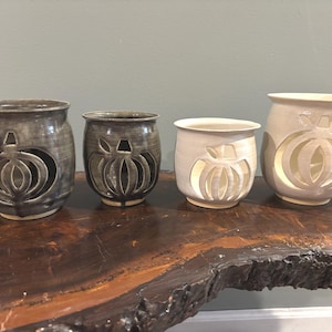 May include: Four ceramic candle holders with pumpkin cutouts. The holders are in shades of gray and white, with a rustic, handmade appearance. They are arranged on a dark wooden surface, perfect for fall decor.