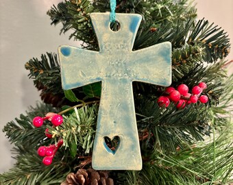 May include: A light blue ceramic cross Christmas ornament with a heart-shaped cutout. The ornament is hanging on a small Christmas tree with red berries and a pine cone. The cross is approximately 4 inches wide.