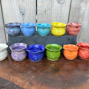 May include: Ten small, handmade ceramic pots in various colors, including white, purple, blue, green, orange, and red. The pots have a scalloped rim and a drainage hole.