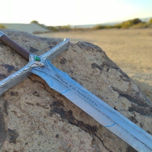 Puede incluir: Una espada plateada con una hoja azul y una gema verde en la empuñadura. La hoja está inscrita con el texto "Dagur völd Kð sviðheiti".