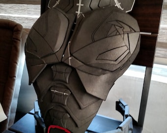 May include: A dark gray, segmented chest armor prop with white accent lines. The armor features a detailed design with a red accent at the bottom and is displayed on a wooden chair. This cosplay item is likely for a superhero or sci-fi costume.