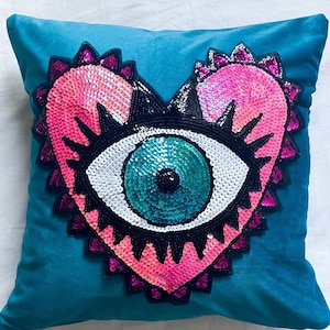 May include: Teal velvet throw pillow with a heart-shaped eye design. The eye is embellished with pink and white sequins, black trim, and a turquoise center, creating a decorative accent for home decor.