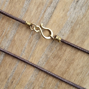 May include: Close-up of a brown cord necklace with a gold-colored hook clasp. The clasp is in the shape of an S-hook. The cord is wrapped with gold-colored wire near the clasp.