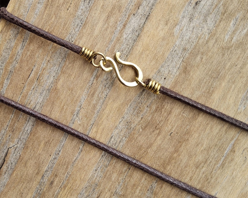 May include: Close-up of a brown cord necklace with a gold-colored hook clasp. The clasp is in the shape of an S-hook. The cord is wrapped with gold-colored wire near the clasp.